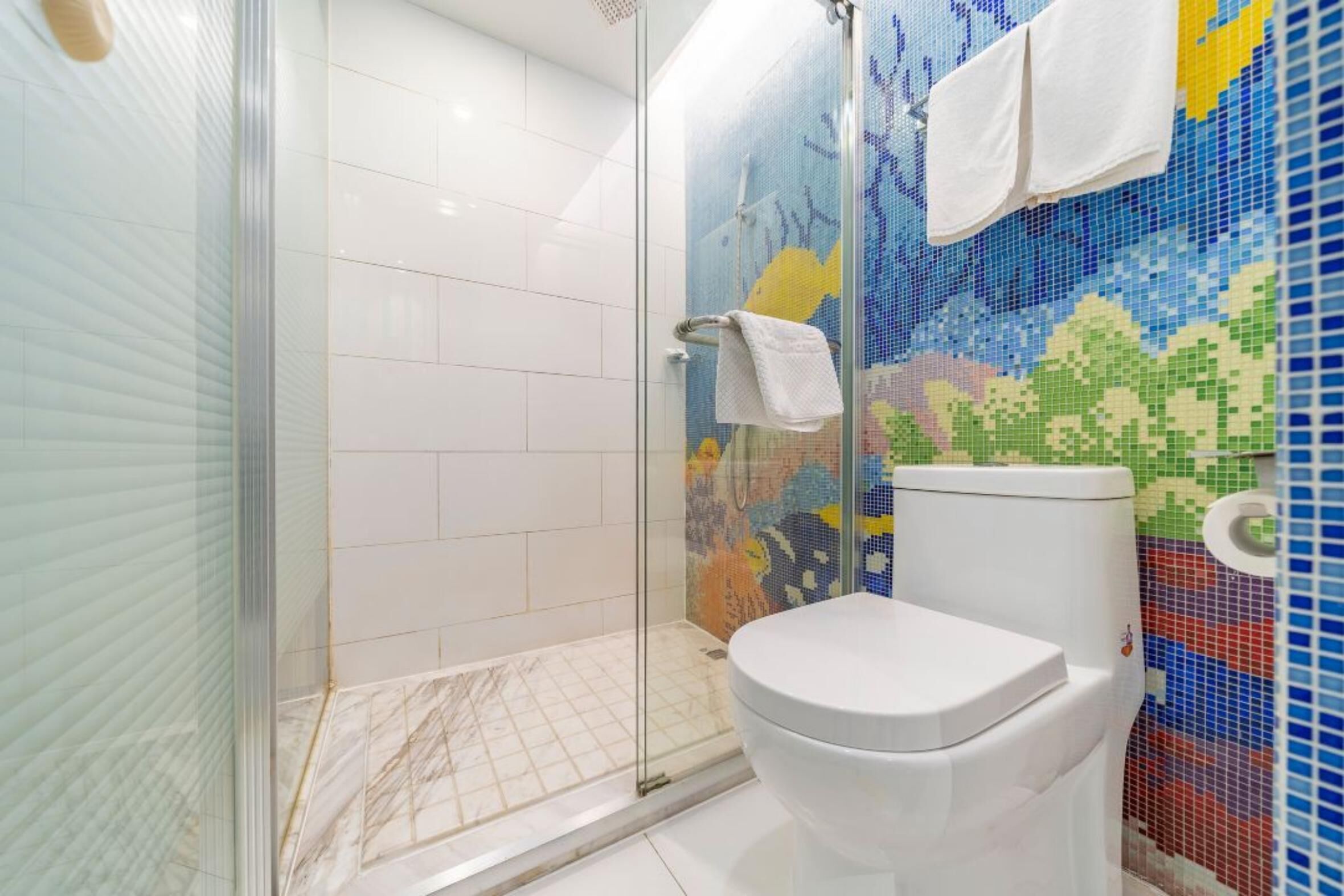 Een badkamer met een douchecabine van glas, een toilet en een wand betegeld met mozaïek.