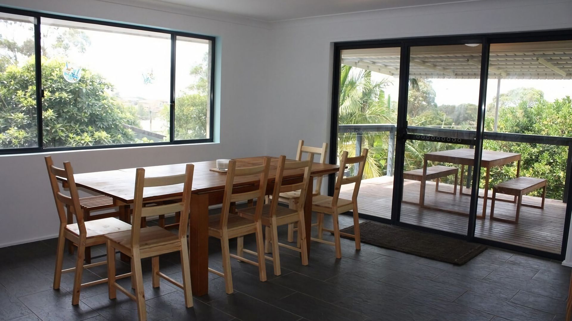 A dining area with a wooden table and chairs, large windows, and a view of greenery.