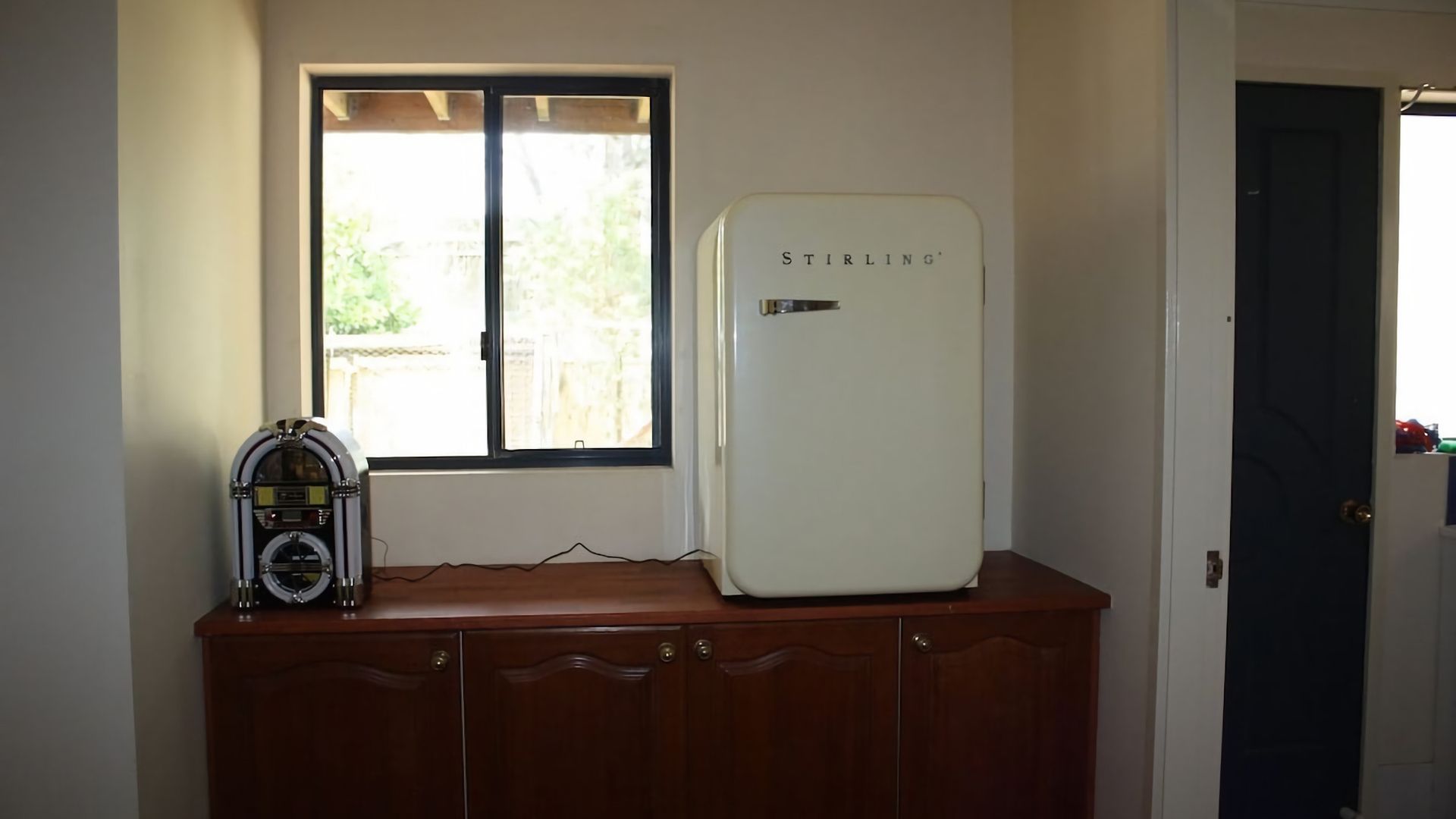 A room with a white refrigerator on a wooden cabinet, a window with a view of greenery, and a door.