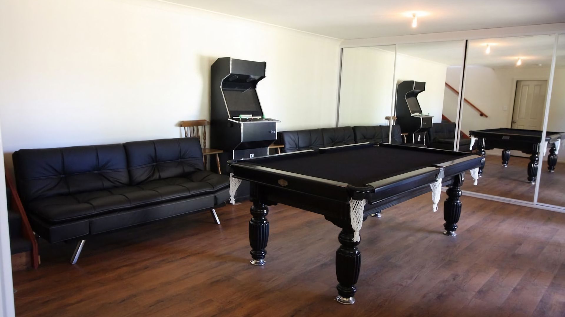 A room with a pool table, a black leather sofa, and a large mirror reflecting the room.