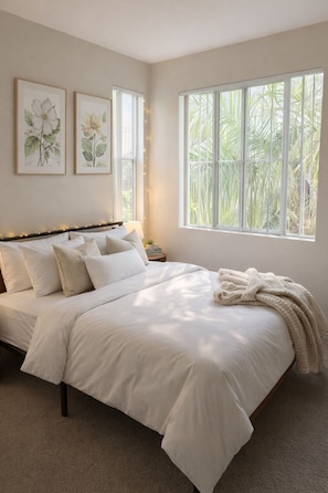 A bedroom with a large bed, two framed botanical prints on the wall, a nightstand, and a window with a view of palm trees. - Stay Demo 1BA Hideaway In Marina Del Rey (Marina del Rey)