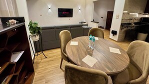 Habitación de hotel moderna con una mesa de comedor de madera, sillas color beige, un sofá gris, una planta pequeña y un televisor de pantalla plana montado en la pared.
