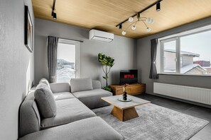 A modern living room with a grey sectional sofa, a wooden coffee table, a TV on a wooden stand, and a large window with a view of buildings. - ASTAGE VILLA KAMIFURANO (Kamifurano)