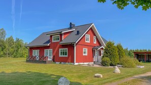 Een rode, twee verdiepingen tellende woning met een schuin dak, een veranda met een leuning en een kleine tuin met rotsen en een pad.