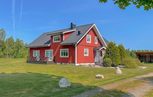 Une maison rouge à deux étages, avec un toit en pente, une véranda dotée d’une balustrade et un petit jardin agrémenté de rochers et d’un sentier.