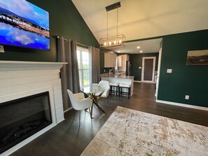 Een moderne woonkamer met een open haard, een eethoek met een glazen tafel, en een keuken op de achtergrond.