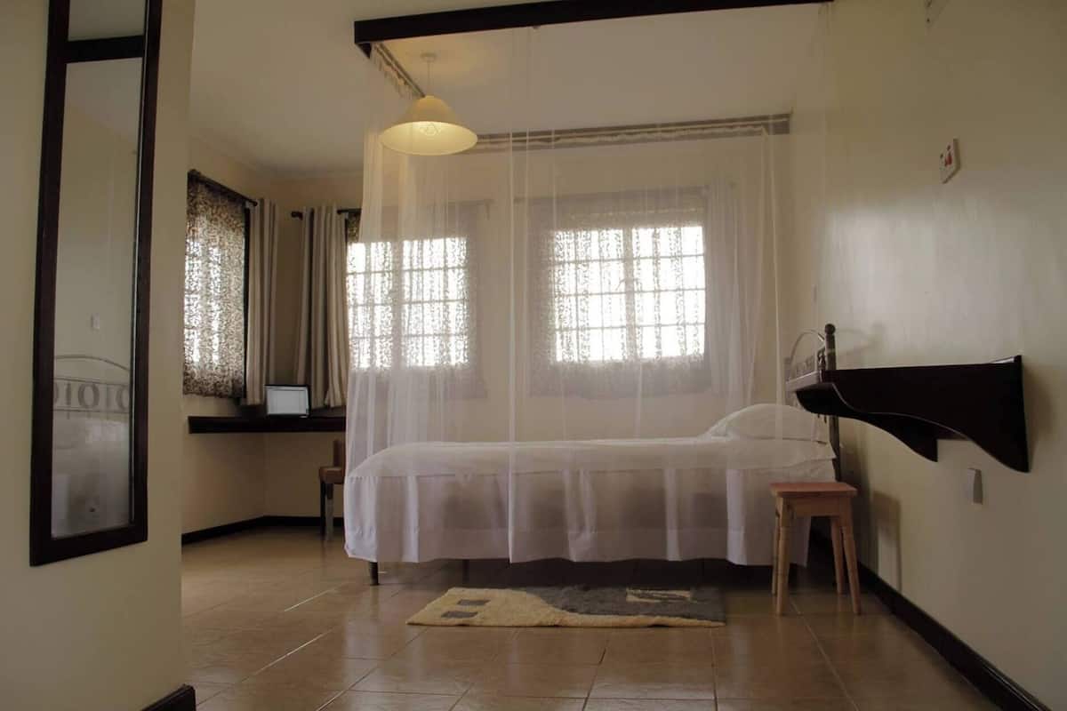 A bedroom with a bed, a bedside table, a nightstand, a window with curtains, and a wall-mounted shelf.