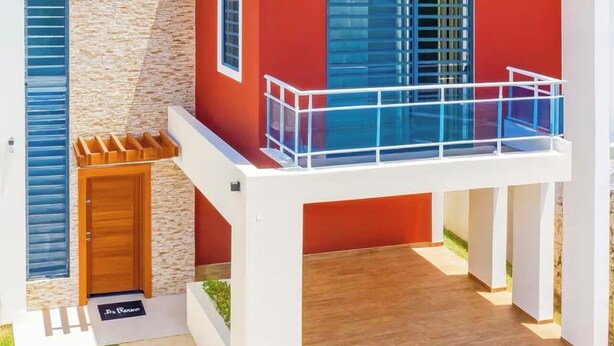 Une maison moderne à deux étages, dotée d’une façade rouge, d’un balcon avec des garde-corps en verre et d’un mur extérieur à l’aspect pierre.
