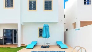 Un espace piscine moderne avec une piscine à l’eau cristalline, deux chaises longues, un parasol bleu et un petit espace dédié à l’exercice physique.