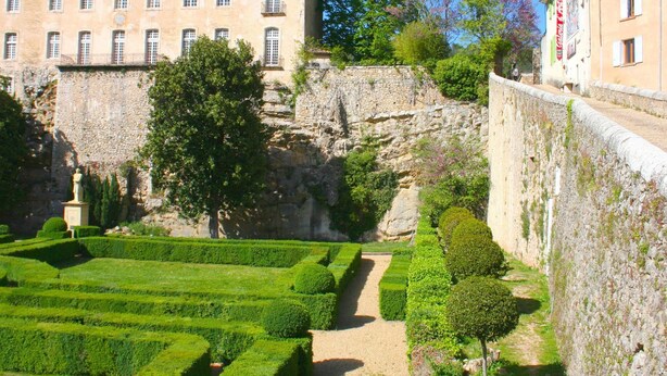Ein Innenhof mit ordentlich geschnittenen Hecken, einer Steinmauer und einem Gebäude mit vielen Fenstern.