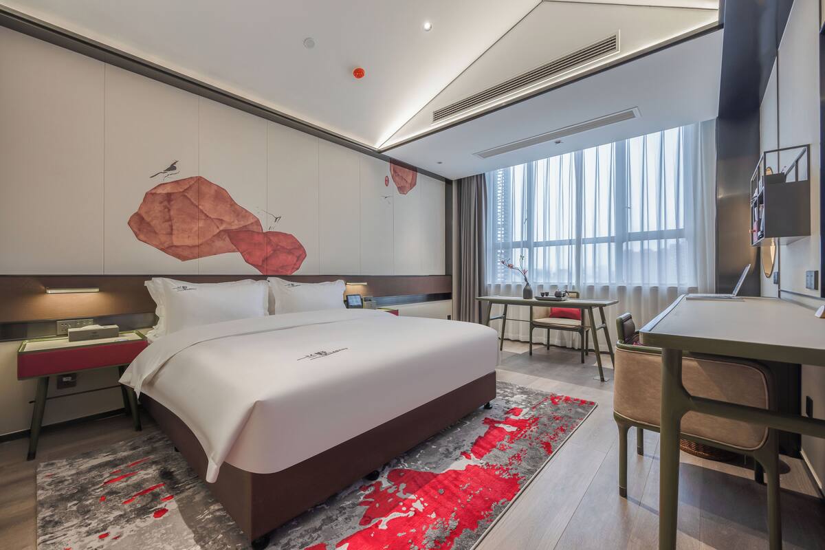 A modern hotel room with a large bed, a desk, and a chair. There is a window with curtains, a wall-mounted TV, and a red abstract artwork.