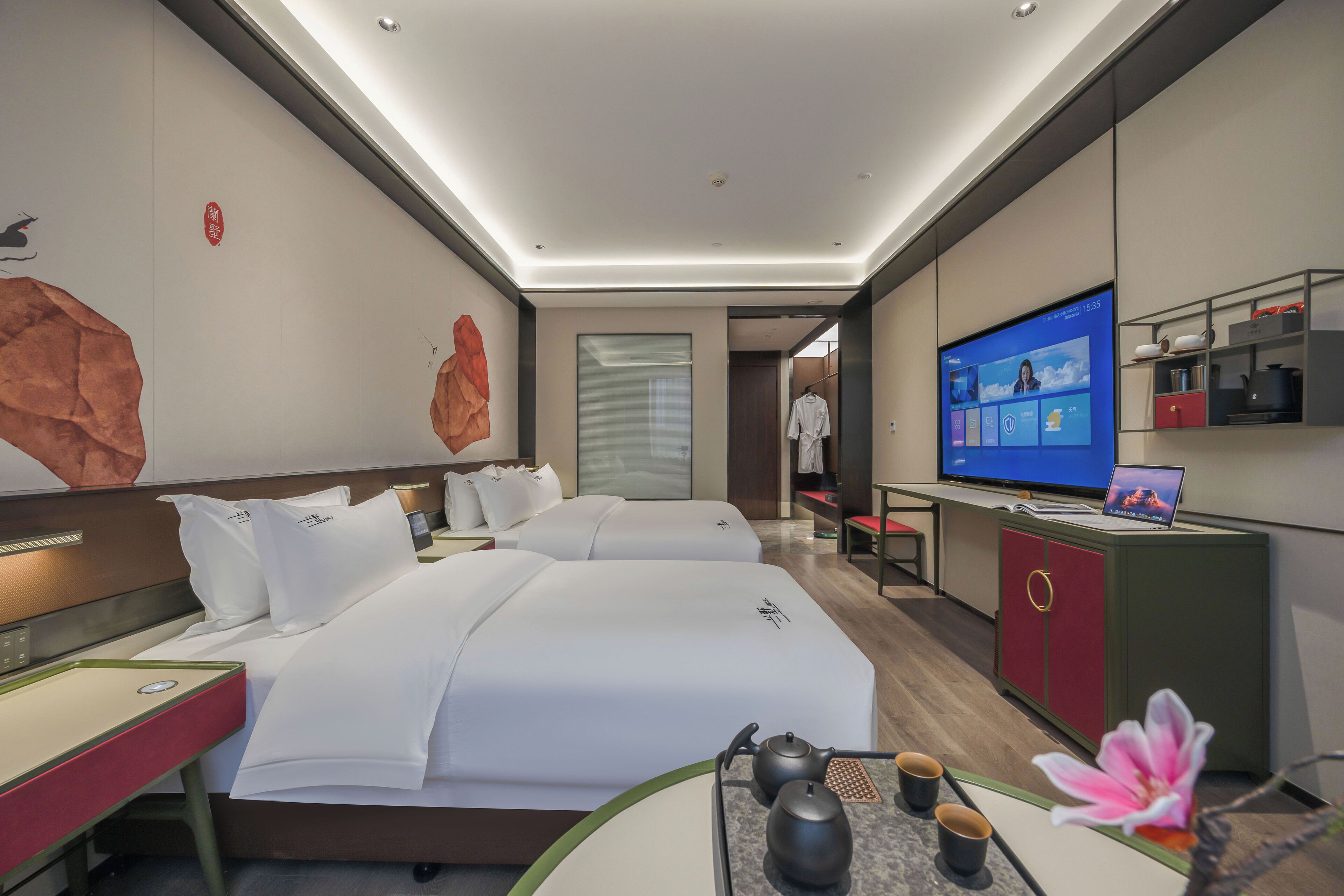 A modern hotel room with two beds, a TV, a desk with a laptop, and a small kitchenette.