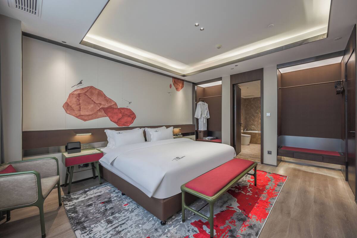 A modern hotel room with a large bed, bedside tables, a bench, a red and grey patterned rug, and a wall art piece.