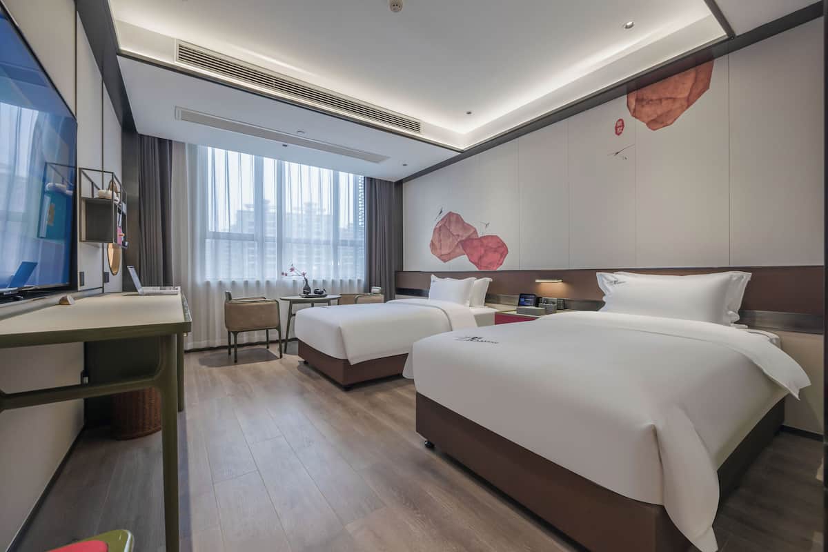 A hotel room with two beds, a desk, a TV, and a large window with a view of buildings.