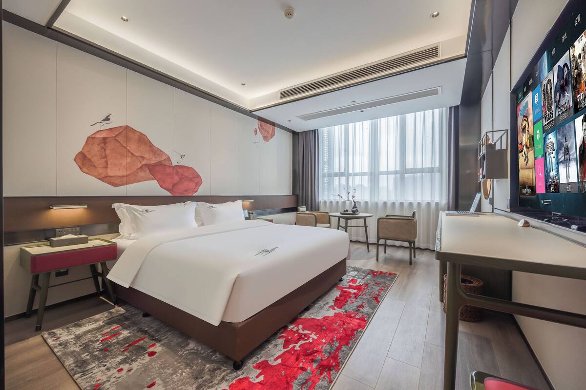 A modern hotel room with a large bed, a desk, a TV, and a view of the city.