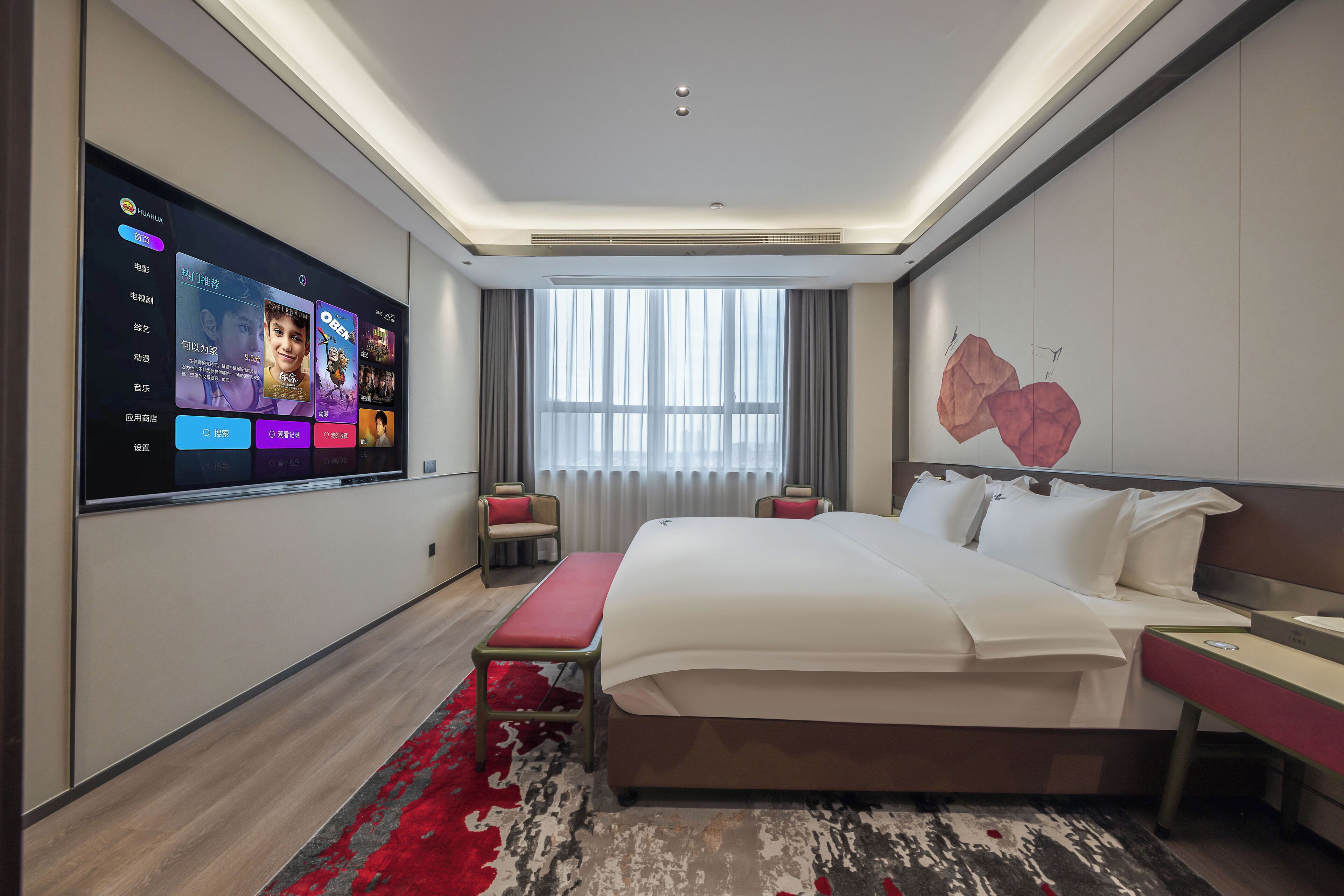 A modern hotel room with a large flat-screen TV on the wall, a large bed with white linens, a red bench, and a red and grey patterned rug.
