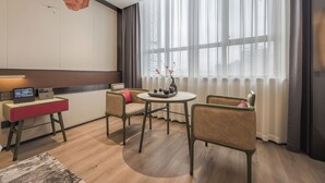 Une chambre d’hôtel moderne avec un plancher en bois, une table ronde entourée de chaises, un bureau avec un ordinateur et une fenêtre ornée de rideaux légers.
