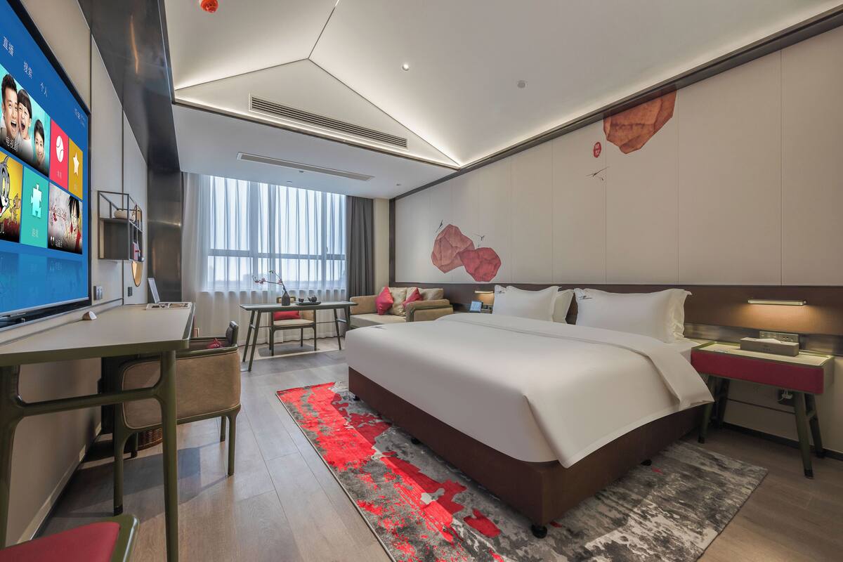 A modern hotel room with a large bed, a desk, and a TV displaying a menu.
