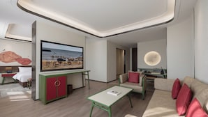 Une chambre d’hôtel moderne dotée d’un grand téléviseur à écran plat, de meubles verts et rouges, d’un canapé avec des coussins rouges et d’un design minimaliste.