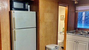 A compact kitchen with a white refrigerator, a microwave on top, a small trash bin, and wooden cabinets. - Cozy Studio Cabin in Kasilof – Comfort Close to Nature (Kasilof)