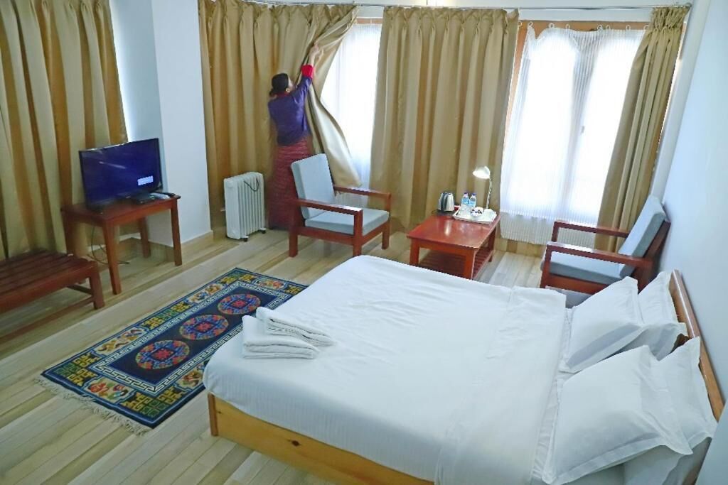 Une chambre d’hôtel avec un lit, deux chaises, une petite table, un téléviseur et une fenêtre avec des rideaux que l’on est en train de régler.