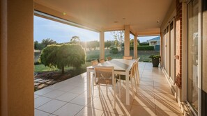 Une terrasse couverte avec une table et des chaises, de grandes fenêtres donnant sur le jardin et les arbres, et une plante en pot posée sur le sol.