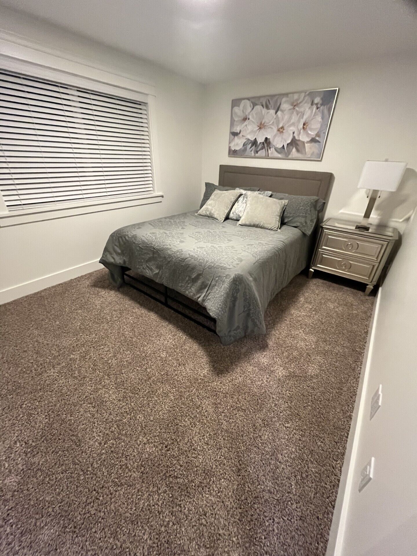 A bedroom with a bed, a nightstand, a lamp, a window with blinds, and a floral painting on the wall.