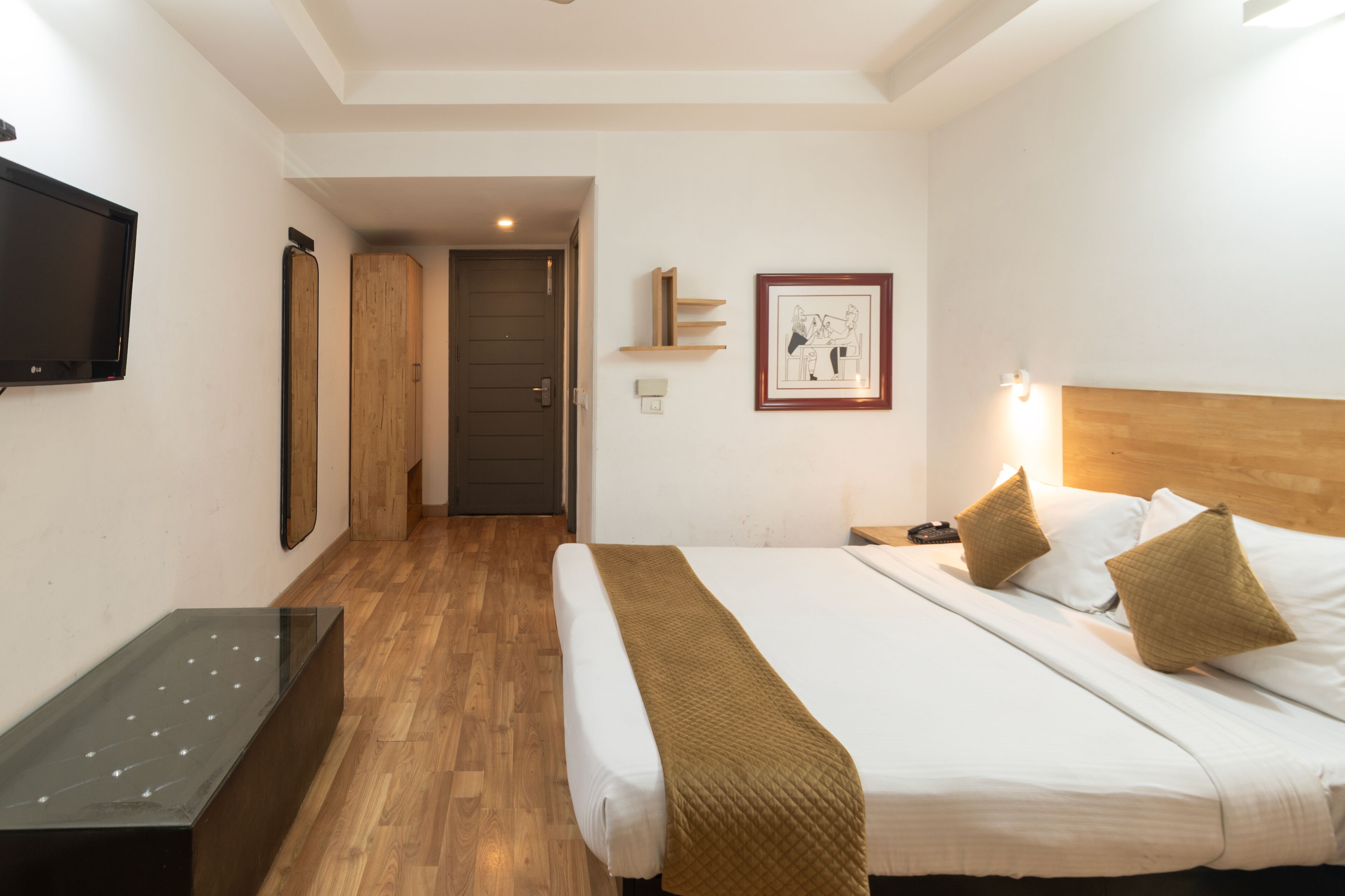 A hotel room with a bed, bedside table, TV, wooden wardrobe, and a framed picture on the wall.