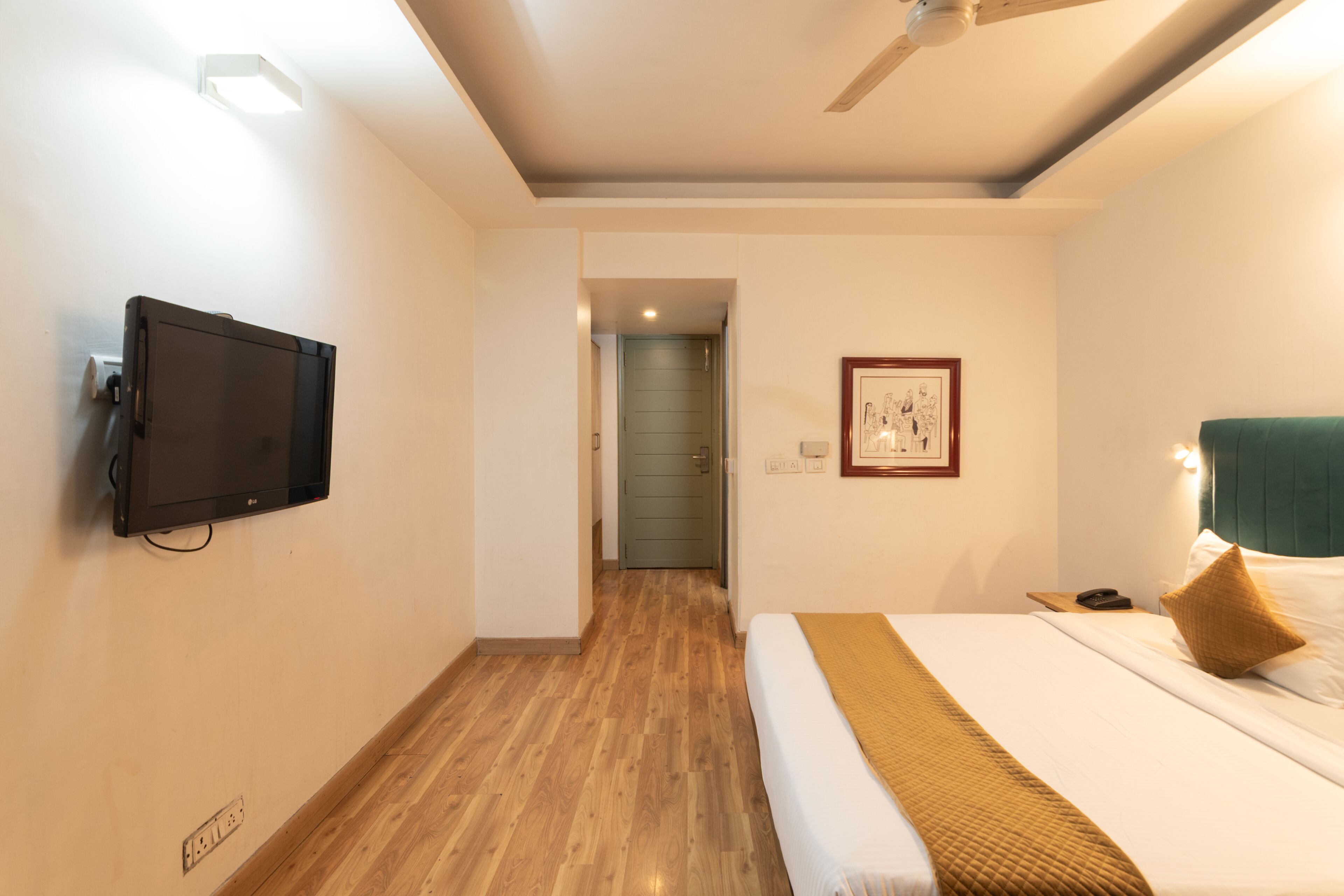 A hotel room with a bed, a television mounted on the wall, a ceiling fan, and a framed picture on the wall.