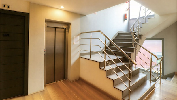 A modern interior with a wooden floor, a staircase with metal railings, and a door with a control panel. - First By JPM (New Delhi)