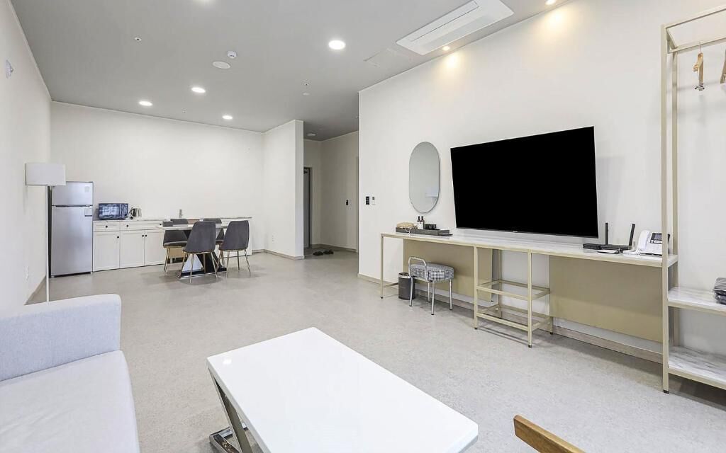 A modern hotel room with a large flat-screen TV, a dining area with a table and chairs, a kitchenette, and a seating area with a sofa and a small table.