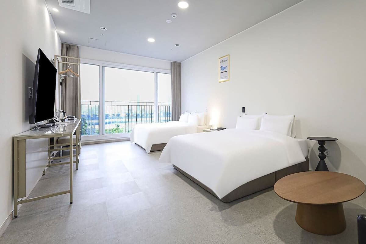 A hotel room with two beds, a desk with a TV, a small table, and a balcony with a view.