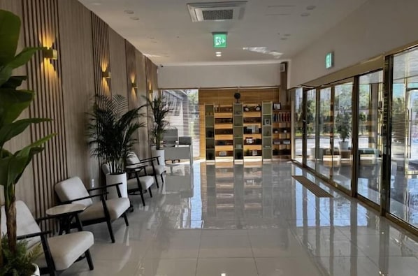 Un vestíbulo moderno con suelo pulido y limpio, zona de estar con sillas y mesas, una estantería de madera y ventanas amplias que ofrecen vistas al exterior.