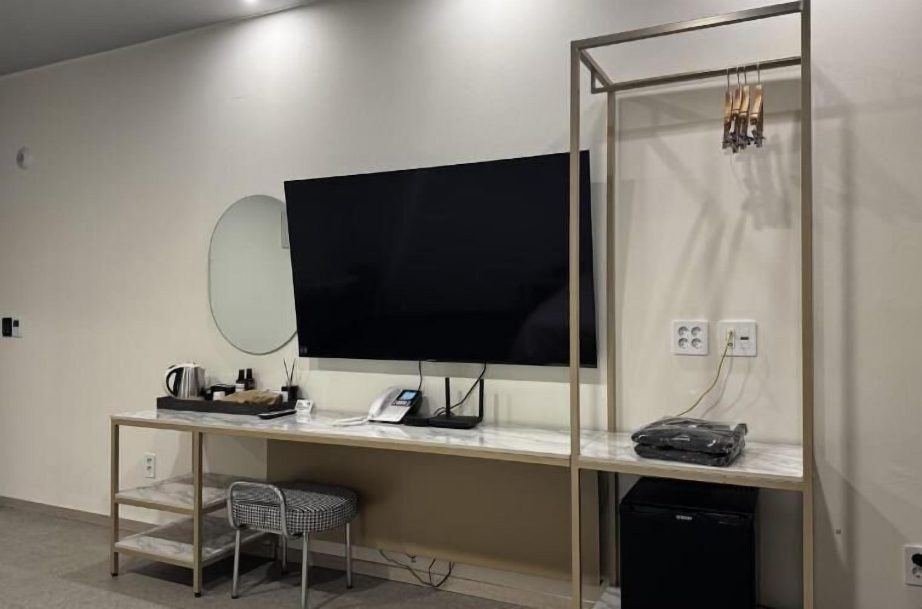 A modern hotel room with a large flat-screen TV mounted on the wall, a desk with a telephone and a power strip, a chair, and a small trash bin.