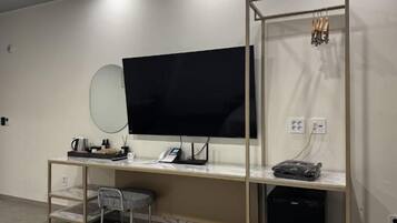 A modern hotel room with a large flat-screen TV mounted on the wall, a desk with a telephone and a power strip, a chair, and a small trash bin.