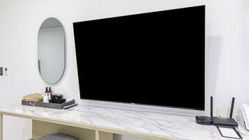 A hotel room with a large flat-screen TV mounted on the wall, a small table with toiletries, a telephone, and a chair.