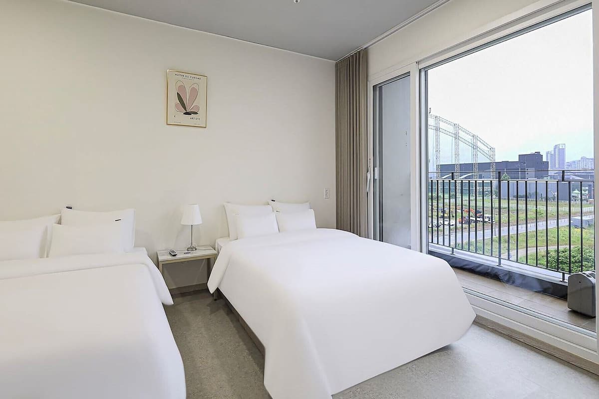 A hotel room with two beds, a bedside table with a lamp, a framed picture on the wall, and a large window with a city view.