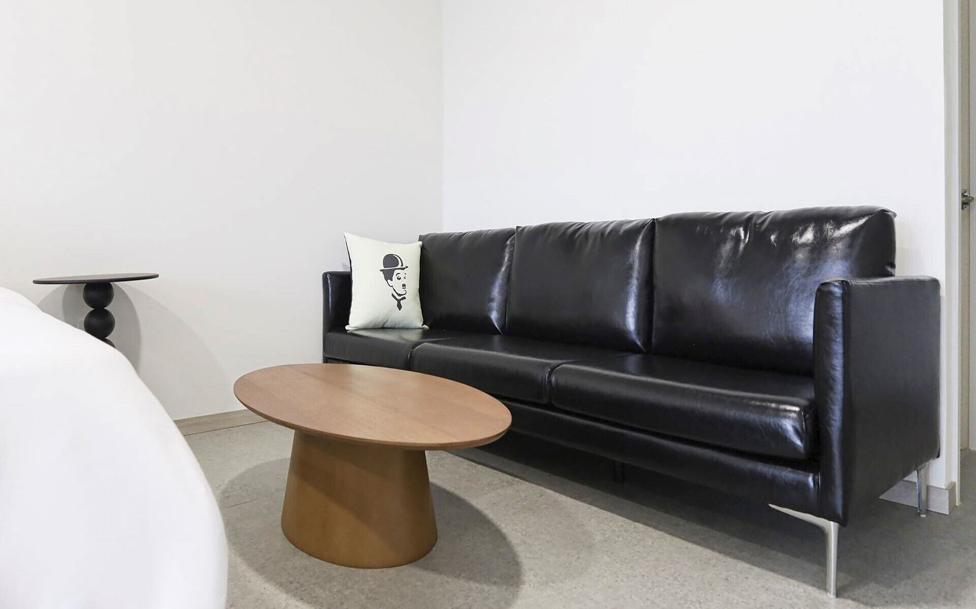 Un sofá de cuero negro con un cojín blanco, una mesa de centro redonda de madera y una mesita negra con tapa redonda.