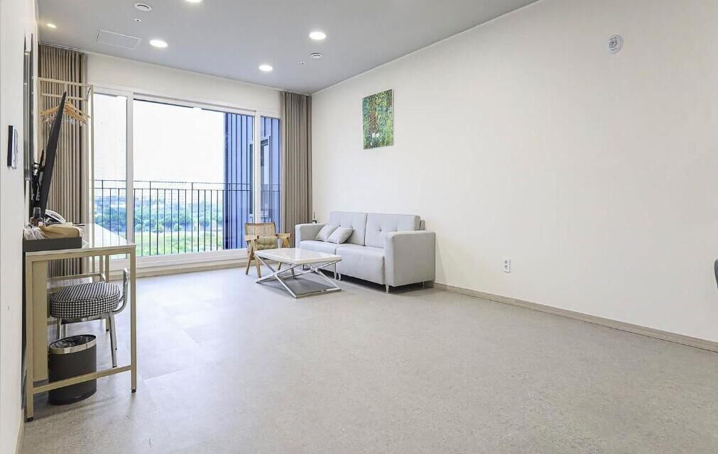 A spacious living room with a white sofa, a small table, and a chair. There is a large window with a view of greenery outside.