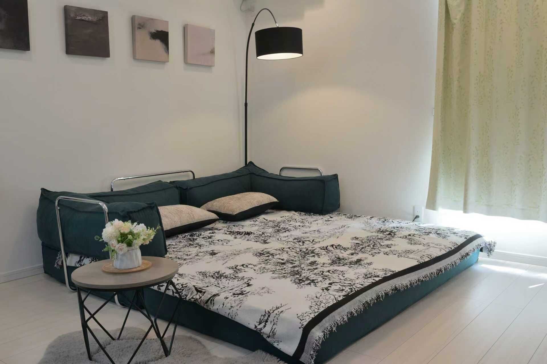 A modern bedroom with a corner bed, a bedside table with a vase of flowers, a floor lamp, and artwork on the wall.
