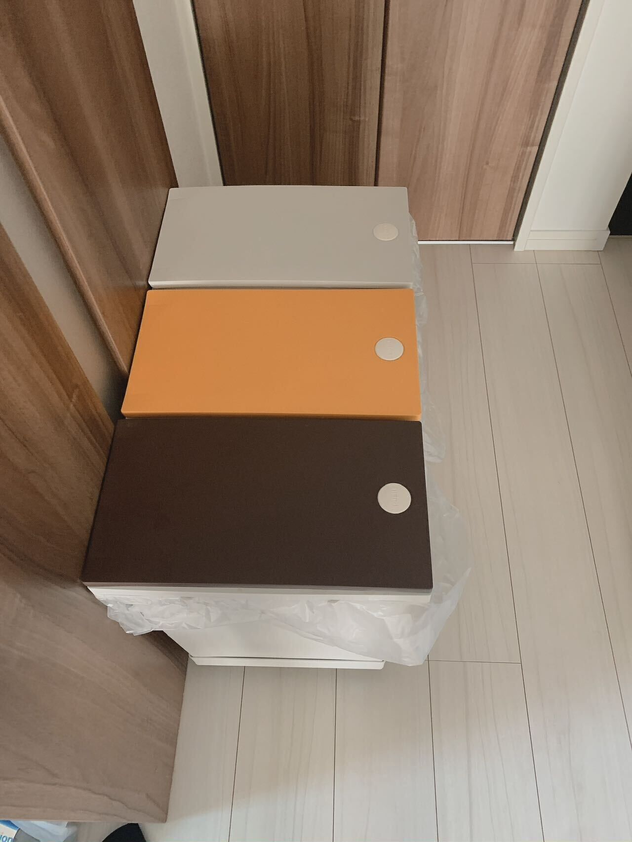 A small, three-tiered, white, brown, and orange storage unit with circular cutouts, placed on a white surface in a room with wooden flooring and walls.