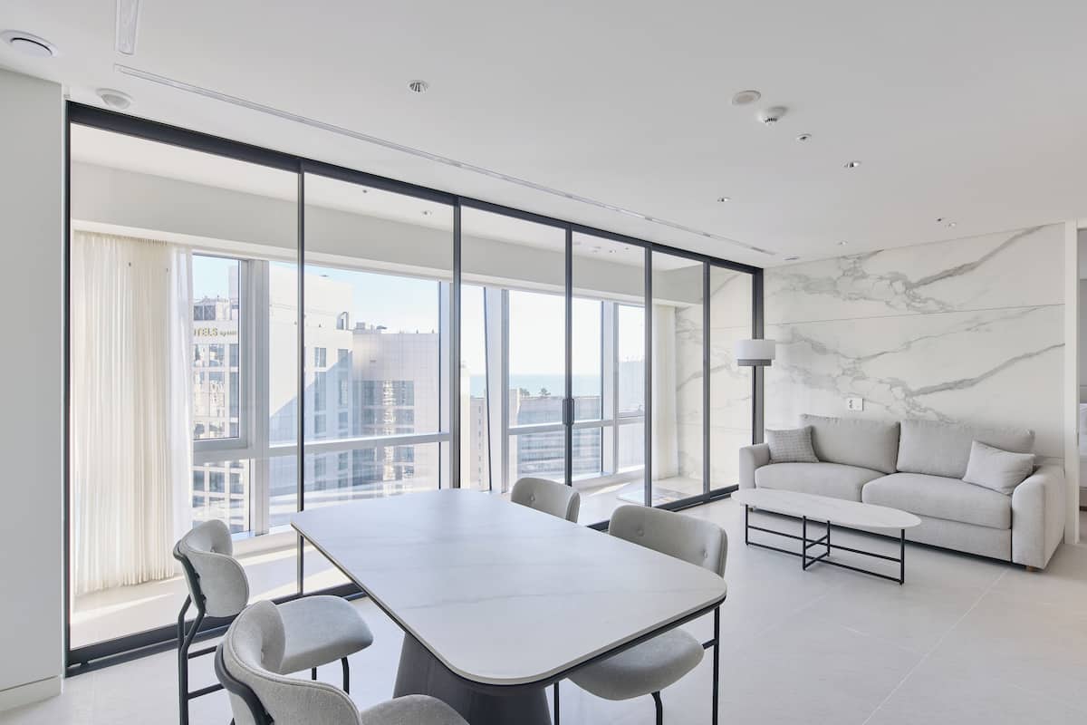 A modern hotel room with a large window, a marble wall, a sofa, and a dining table.