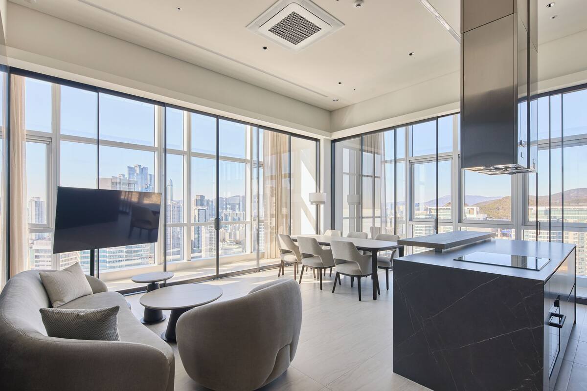 A modern hotel room with a large window, a flat-screen TV, a dining area, and a kitchen island.