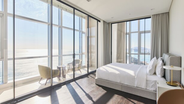 Ein modernes Hotelzimmer mit einem großen Bett, einer Sitzecke mit zwei Sesseln und Meerblick durch raumhohe Fenster.