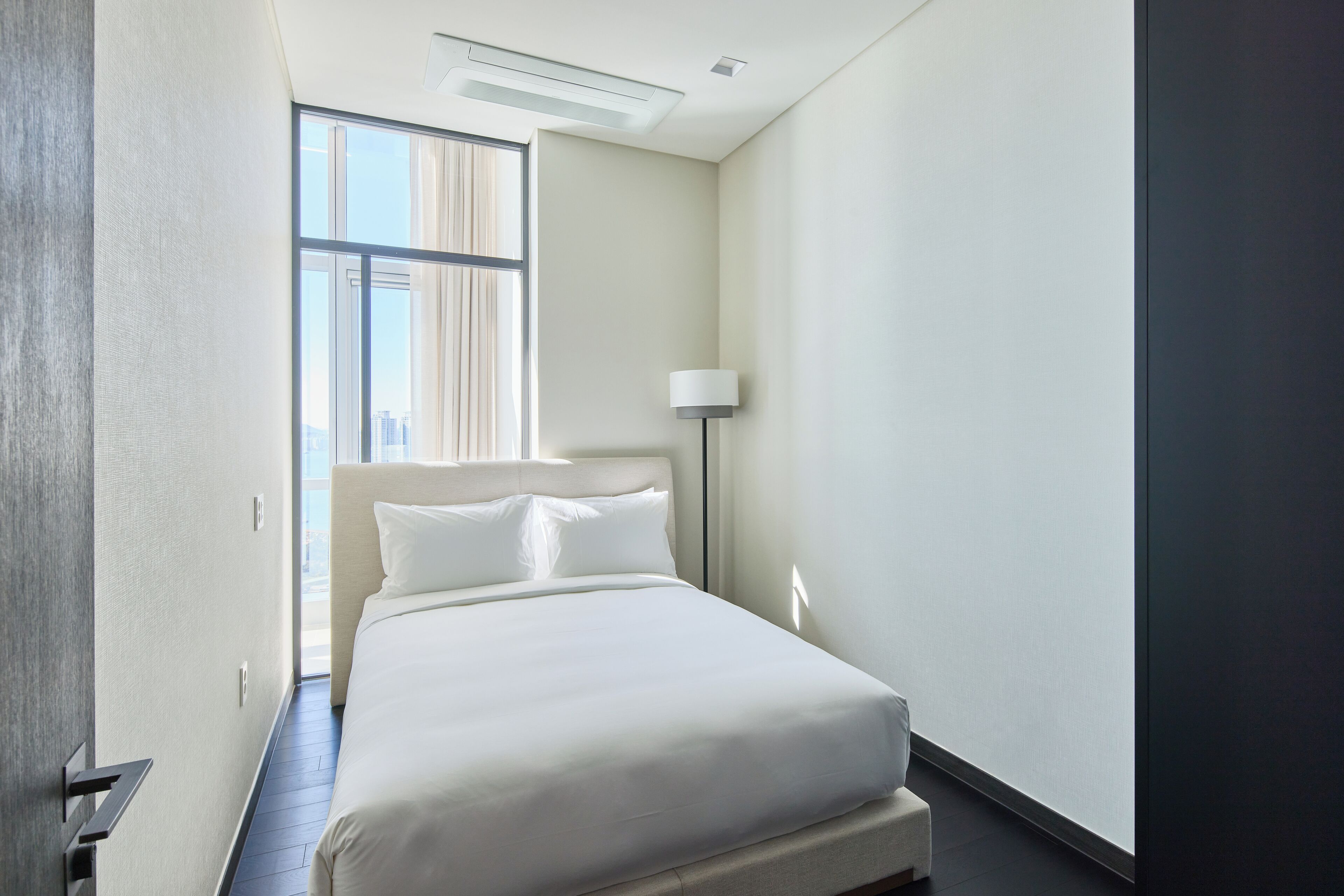 A hotel room with a large bed, a bedside lamp, and a view of the city through a window.