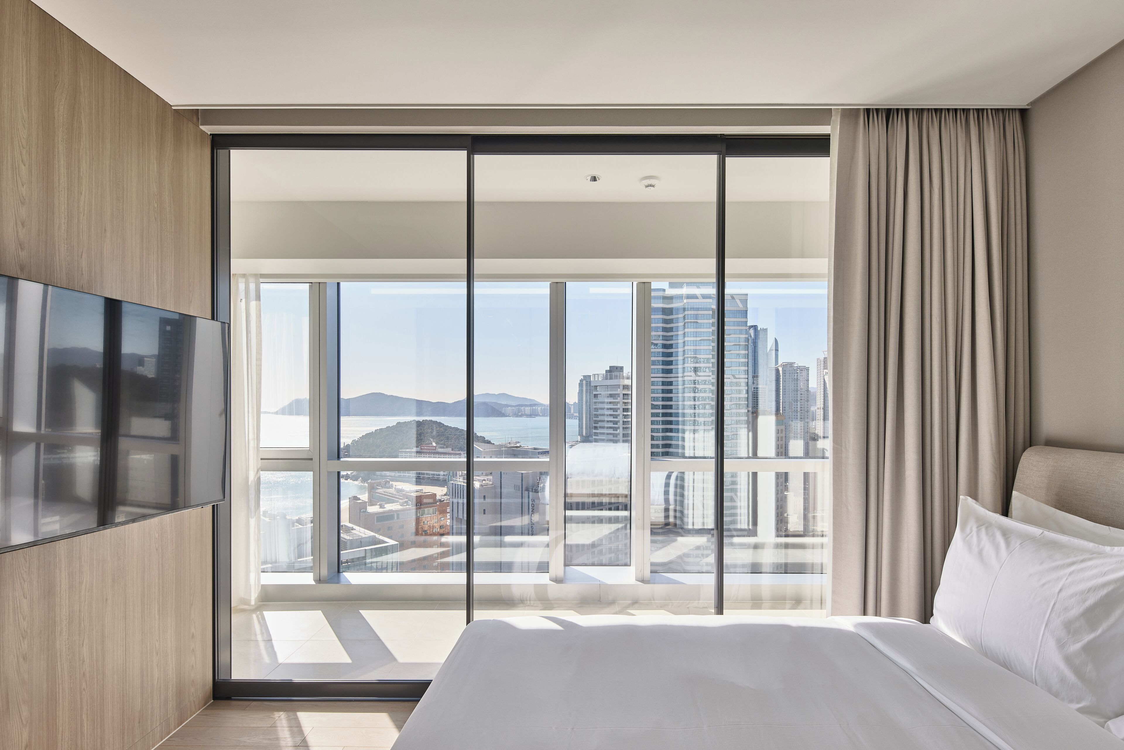 Ein modernes Hotelzimmer mit einem großen Bett, einem flachbildfernseher an der Wand und einem großen Fenster mit Blick auf die Stadt.