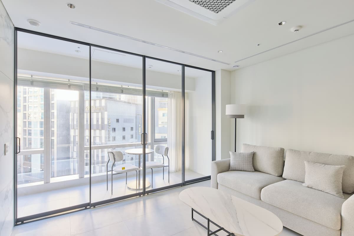A modern hotel room with a large sliding glass door leading to a balcony with a city view. There is a sofa and a small table with chairs on the balcony.