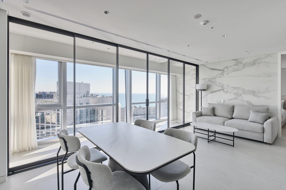A modern hotel room with a large window, a sofa, and a dining table.