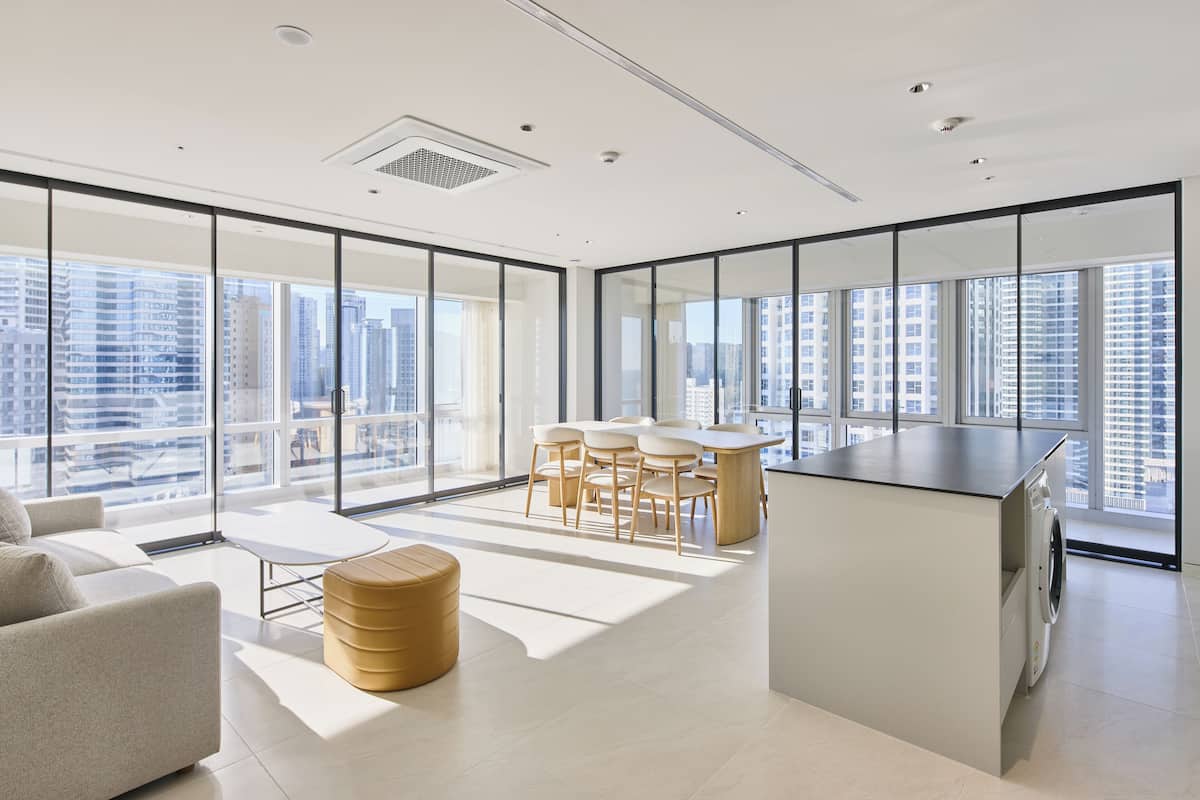A modern hotel room with a large window, a sofa, a coffee table, a dining area with chairs, and a washing machine.