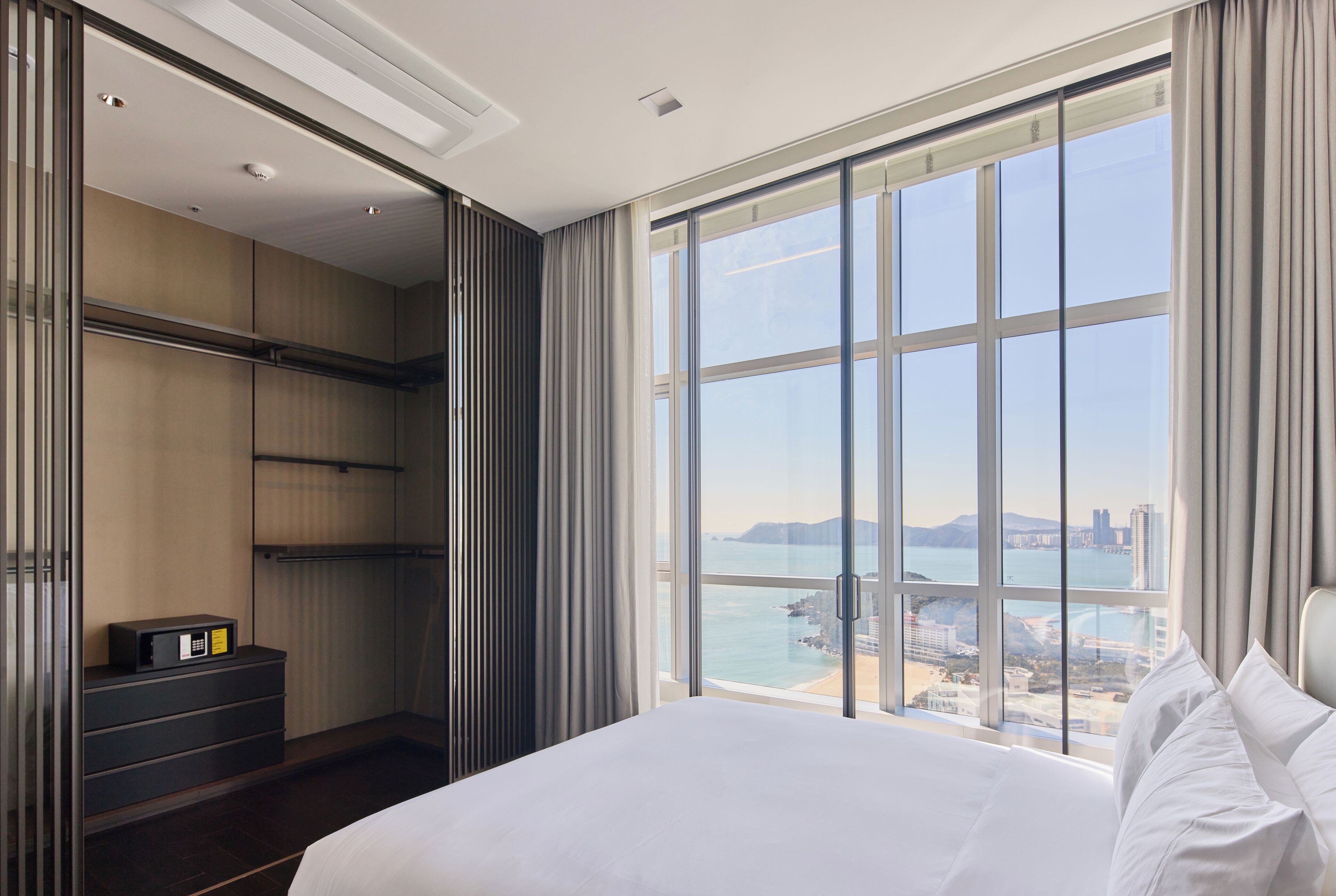 Quarto de hotel com uma cama grande, um guarda-roupa, uma mesa com um computador e vista para o mar e edifícios.
